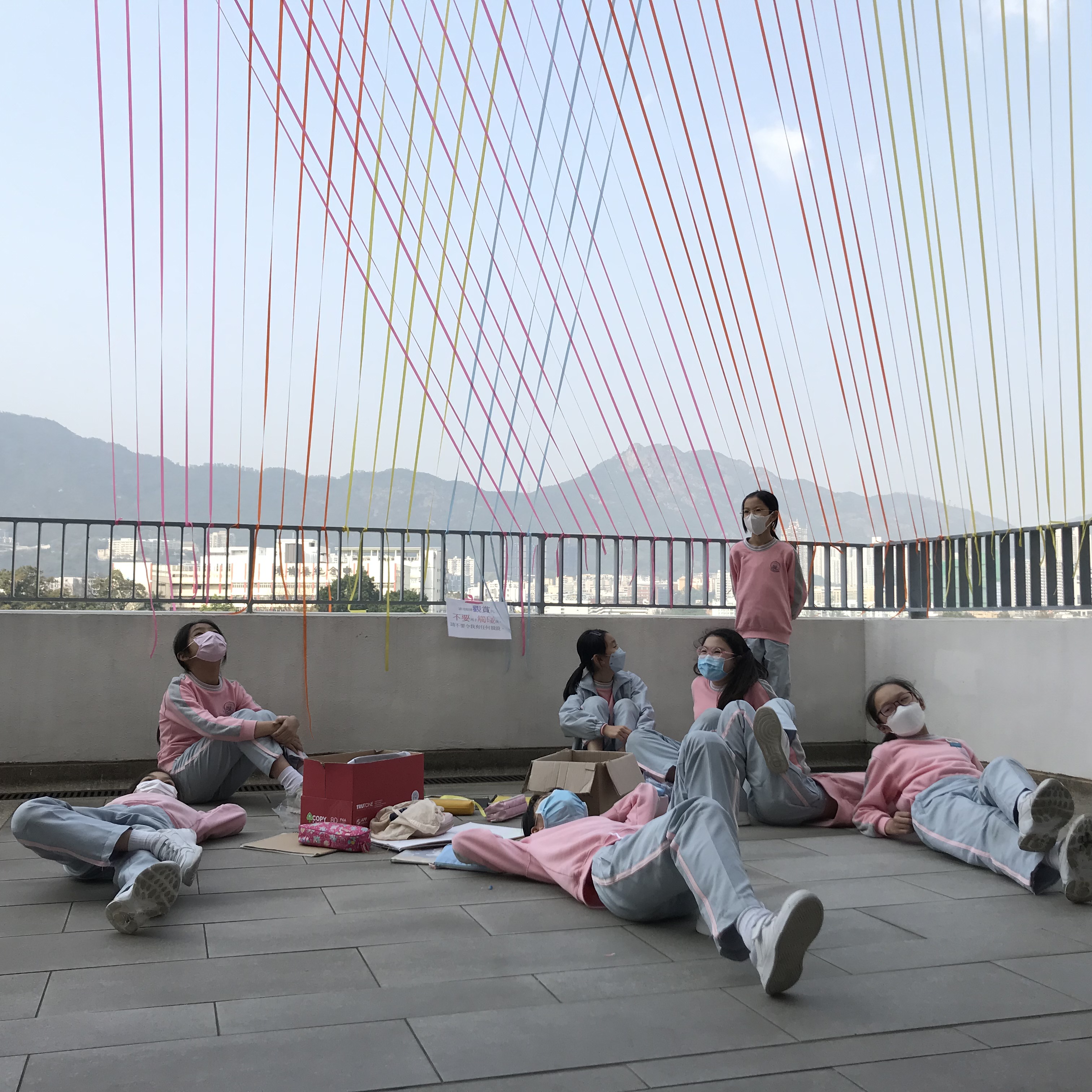 A group of students lying on the ground of a concrete balcony, looking up and there are colourful ropes coming from the ceiling down to the balcony ledge.
